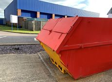 Red industrial dumpster on gravel beside a road, with a blue warehouse in the background