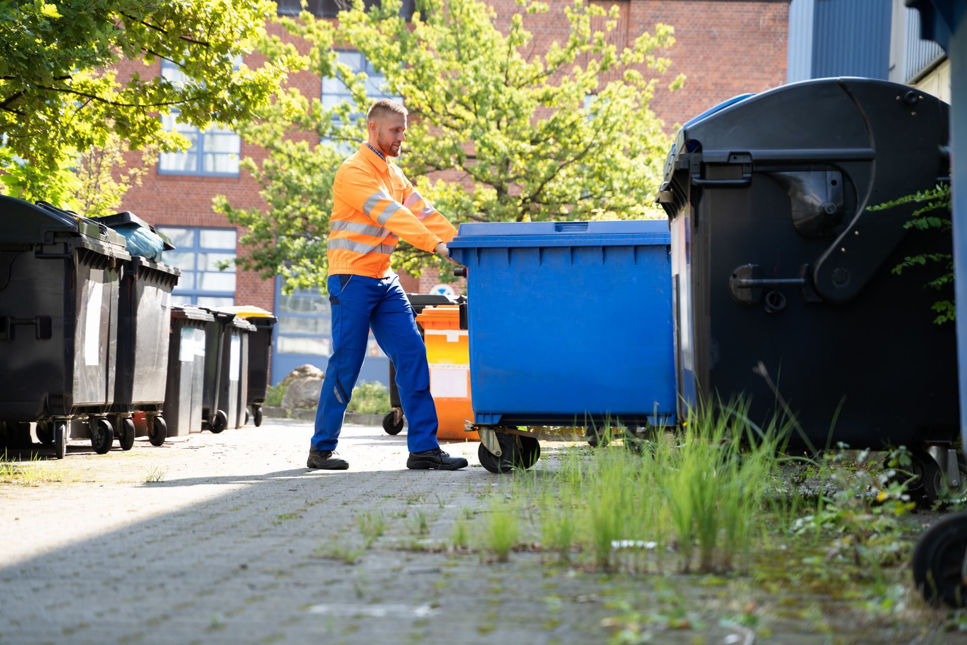 Worker in orange safety jacket moving a blue dumpster along a sidewalk with black bins nearby.
