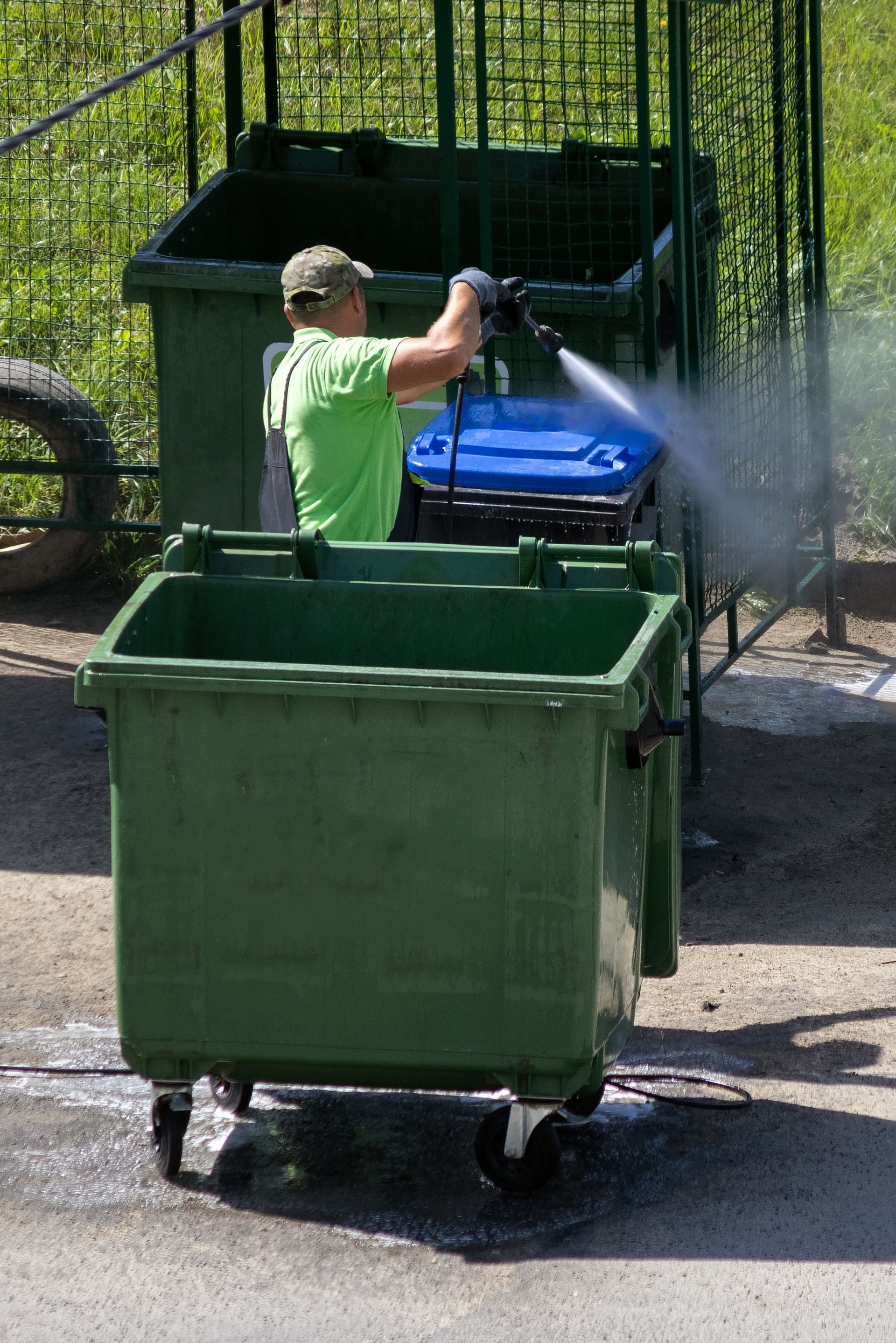 Person pressure-washing blue recycling bins beside a green dumpster outdoors.
