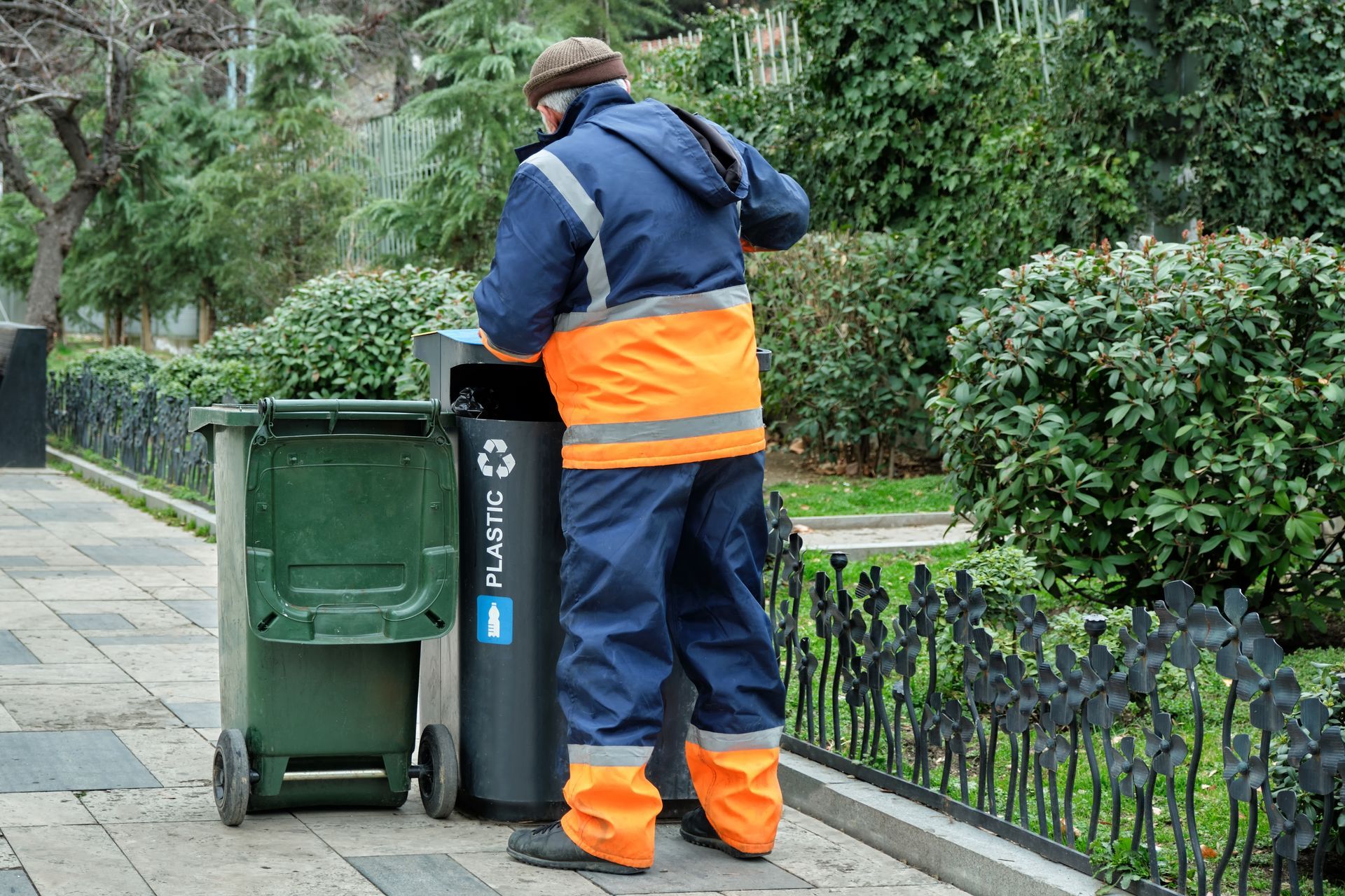 Worker in orange safety gear emptying trash bins on a paved park path, surrounded by greenery.