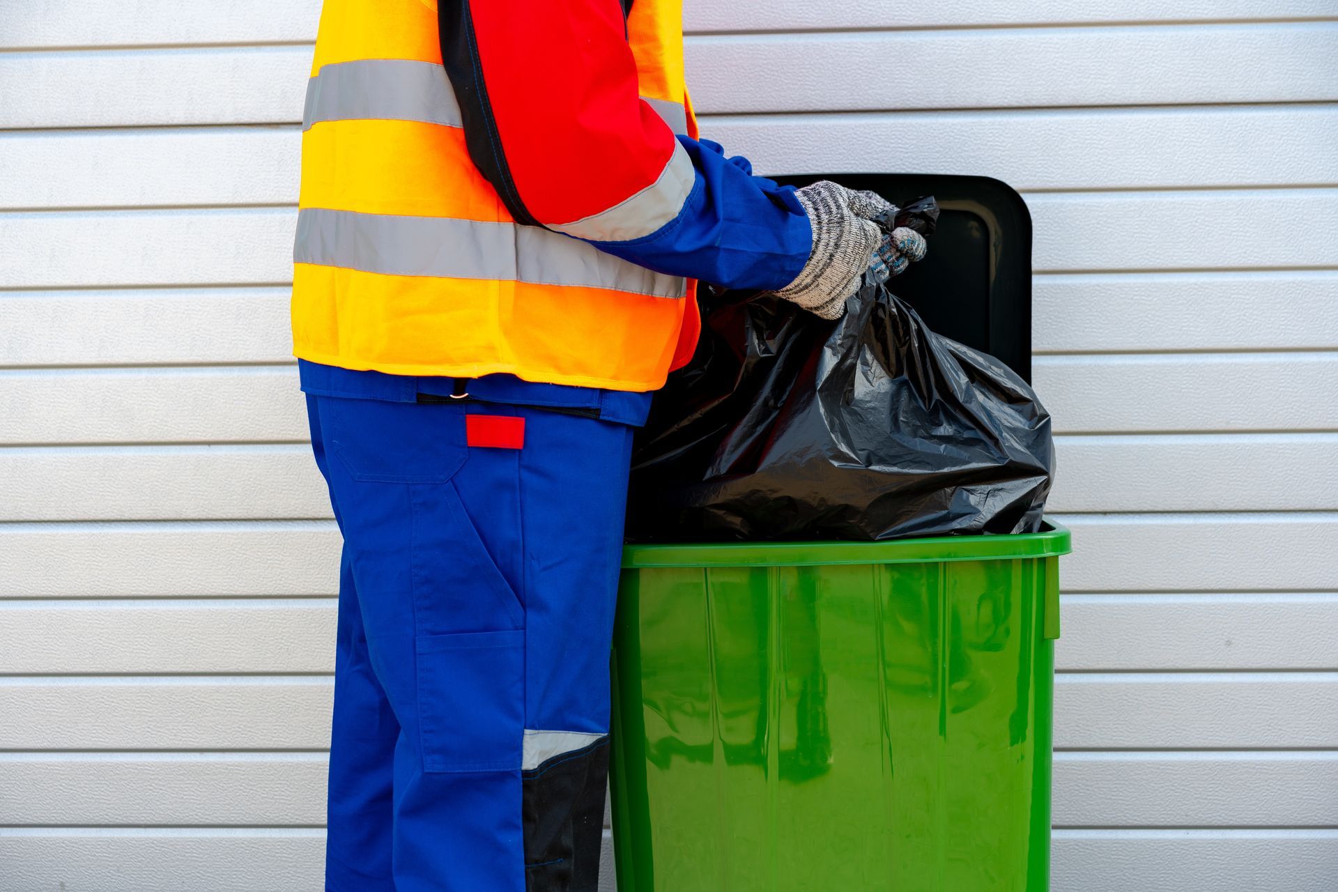 Worker in a reflective vest puts a black trash bag into a green bin.
