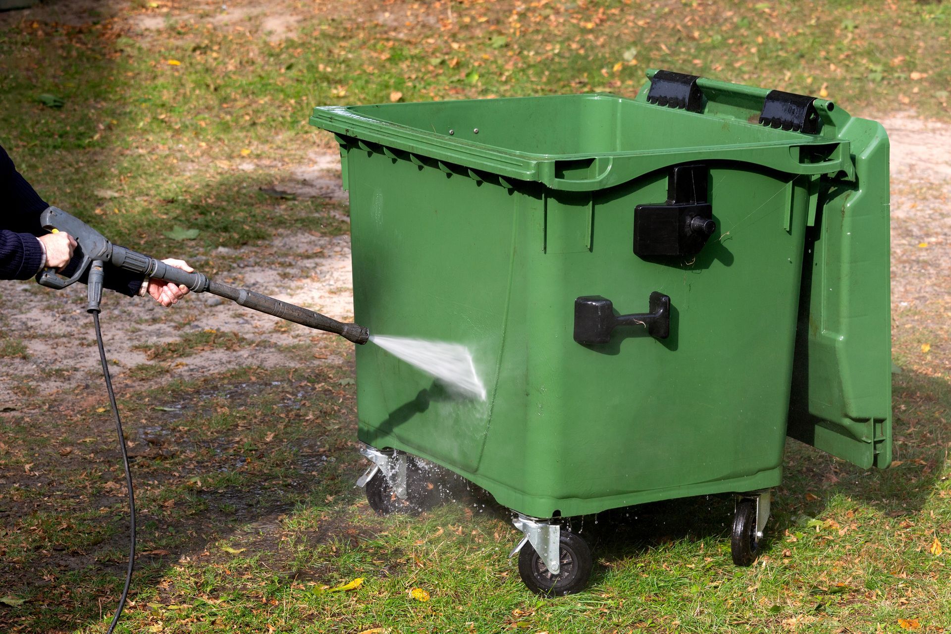 Person pressure-washes a green dumpster outdoors on grass.