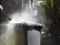 Person pressure-washes a gray trash bin outdoors with a spray of water.