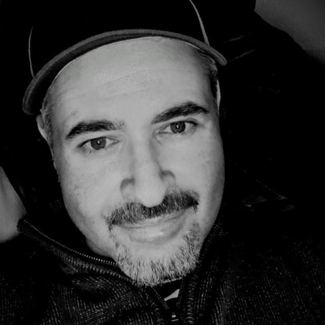 Mann in Schwarz und Weiß, mit Mütze und Jacke mit Reißverschluss, leicht lächelnd.