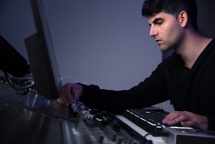 Mann in dunkler Kleidung in einem Musikstudio, der ein Keyboard und andere Geräte an einem Computermonitor bedient.
