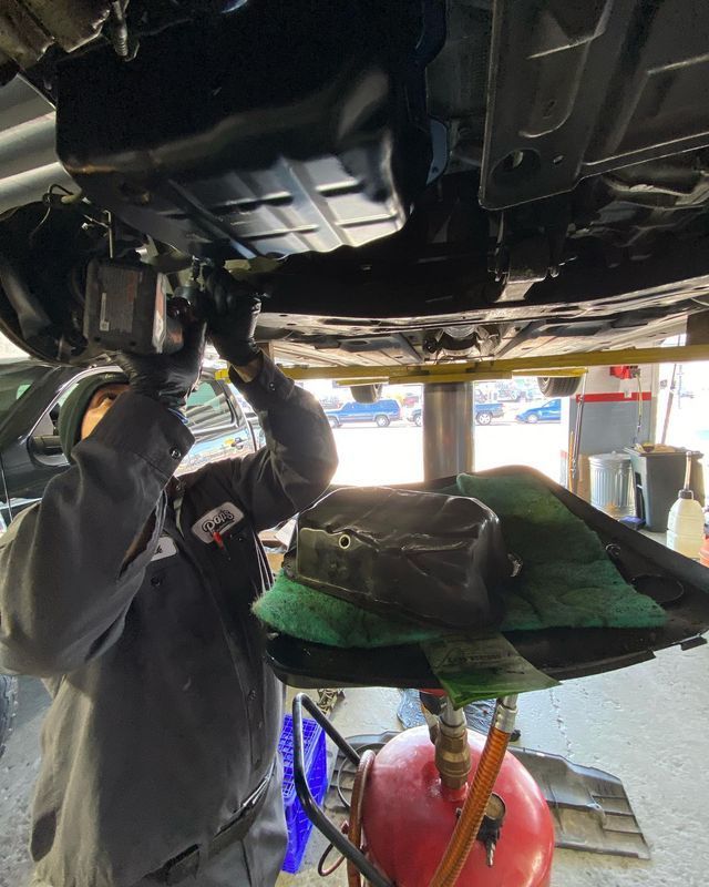 Mechanic under a car draining oil; red oil tank below, green mat and filter on ground.