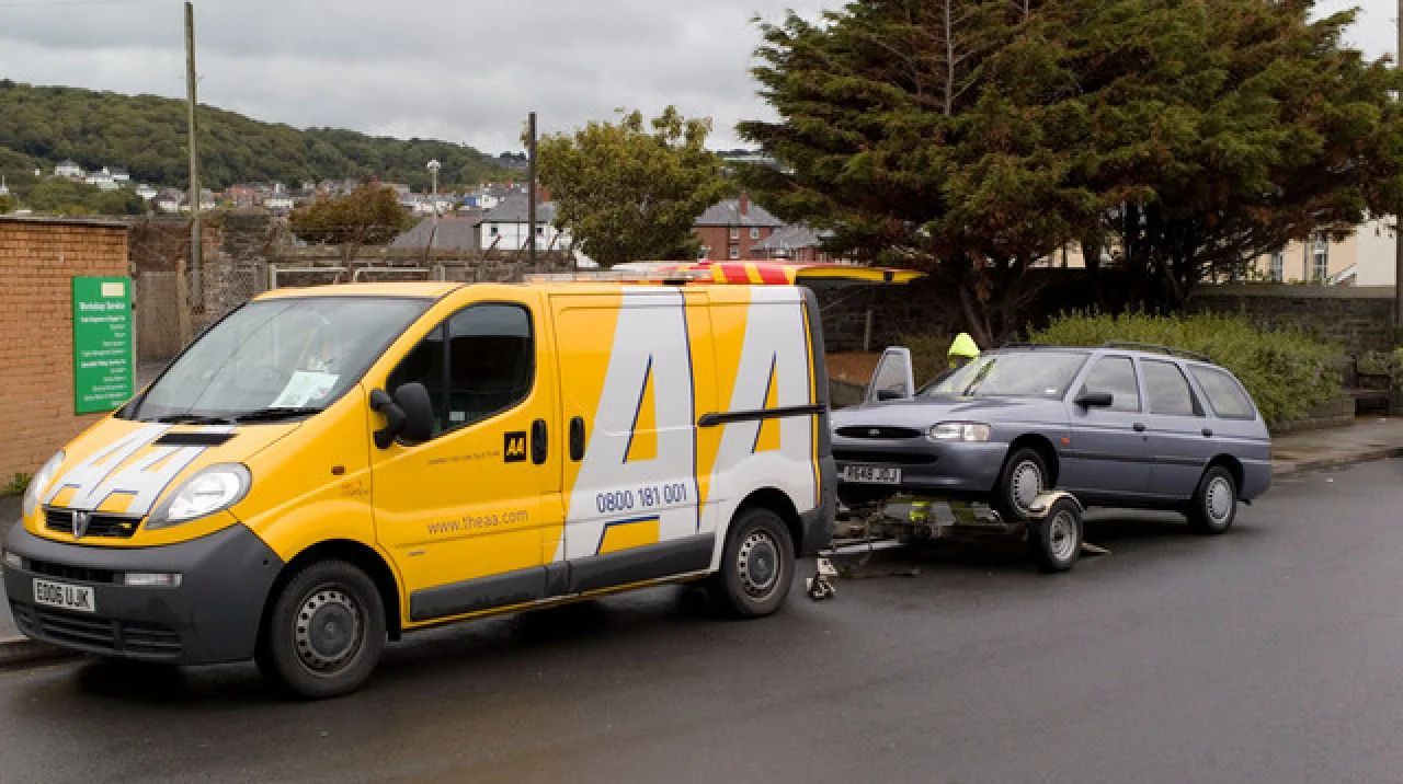 Yellow tow truck towing a gray sedan on a wet roadside.