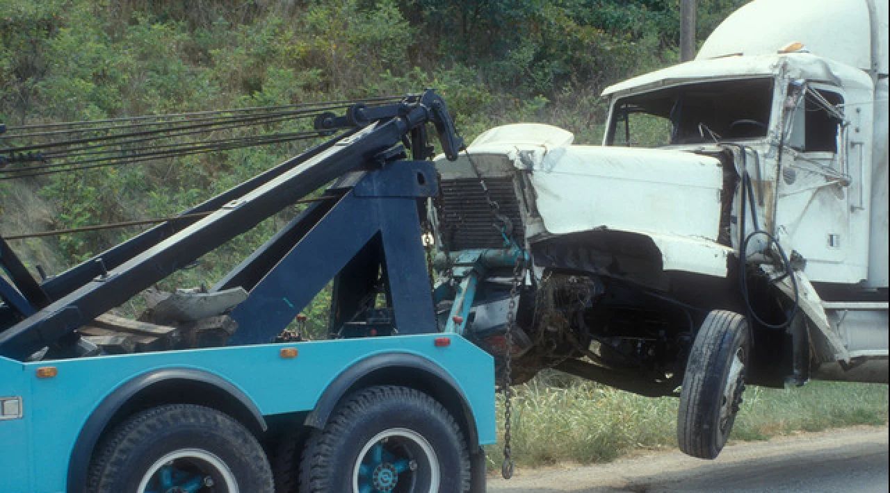Tow truck lifting a white semi-truck off a roadside ditch beside a wooded embankment