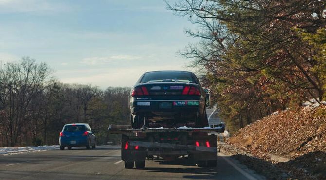 Car carrier truck hauling a black sedan on a highway beside another car and bare trees