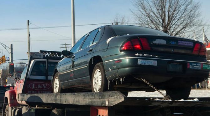 Dark sedan on a tow truck, with another vehicle behind it under a clear sky.