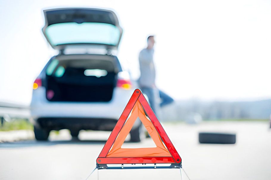 Red warning triangle on roadside in front of a broken-down car with open trunk and a person standing nearby.