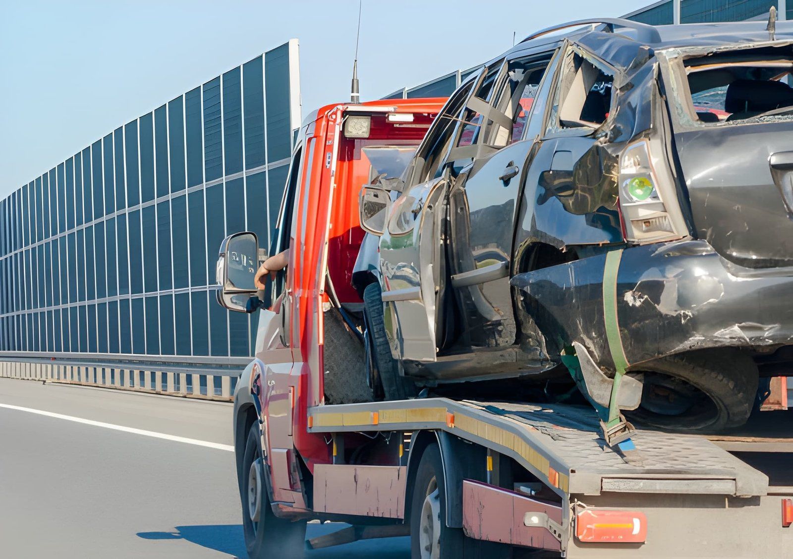 Tow truck carrying a badly damaged black car on a highway beside a blue sound barrier