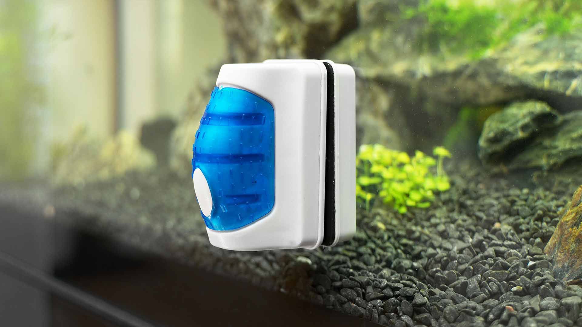 White and blue magnetic aquarium cleaner attached to a glass wall with gravel and plants in the background.