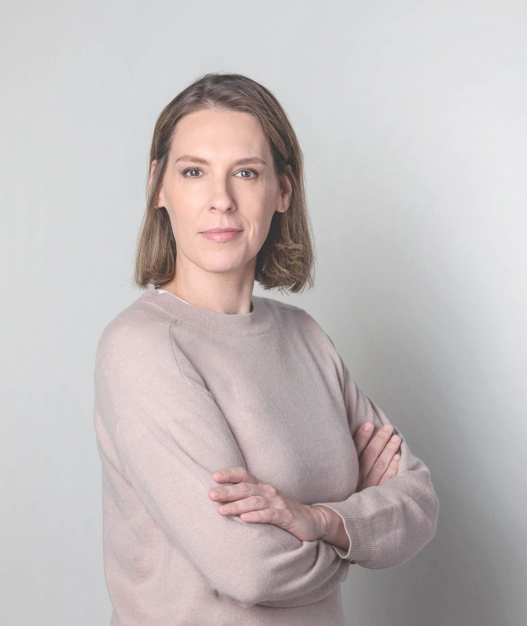 Frau mit verschränkten Armen, hellrosa Pullover, blickt vor grauem Hintergrund in die Kamera.
