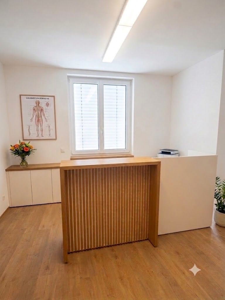 Physiotherapieraum mit Untersuchungstisch, Schreibtisch, anatomischen Diagrammen und zwei grünen Stühlen.