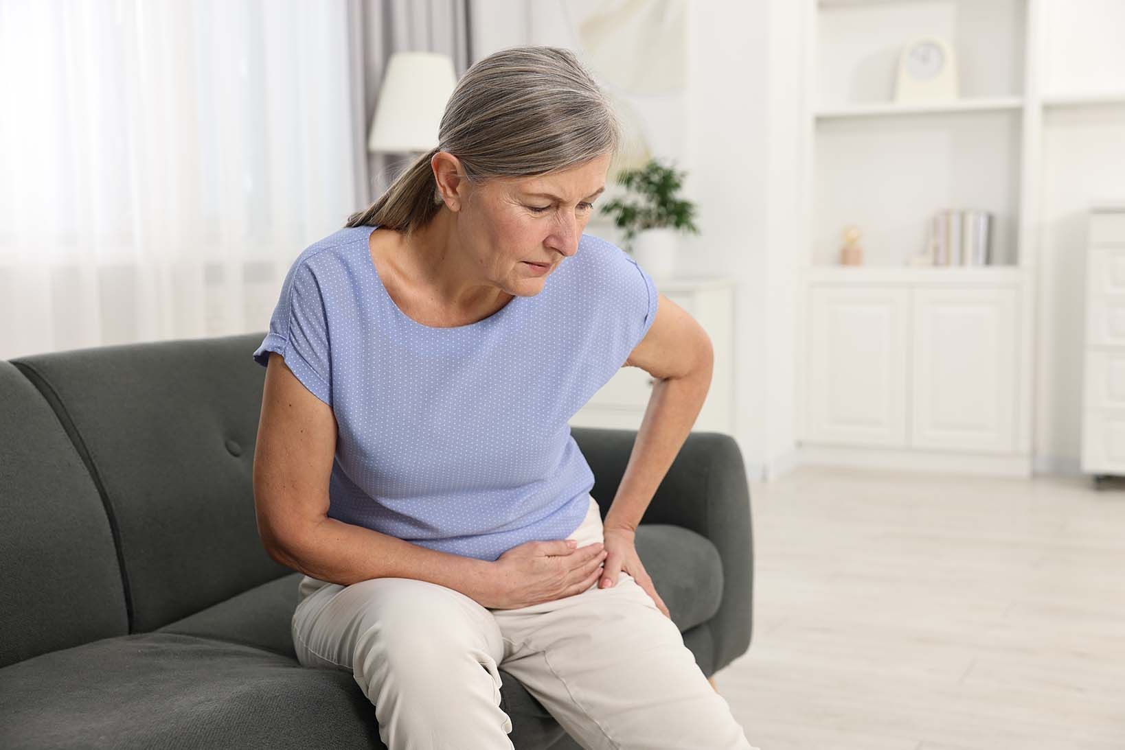 Ältere Dame sitzt auf einemdunelgrauem Sofa und hält Ihre Hände zur schmerzenden Hüfte