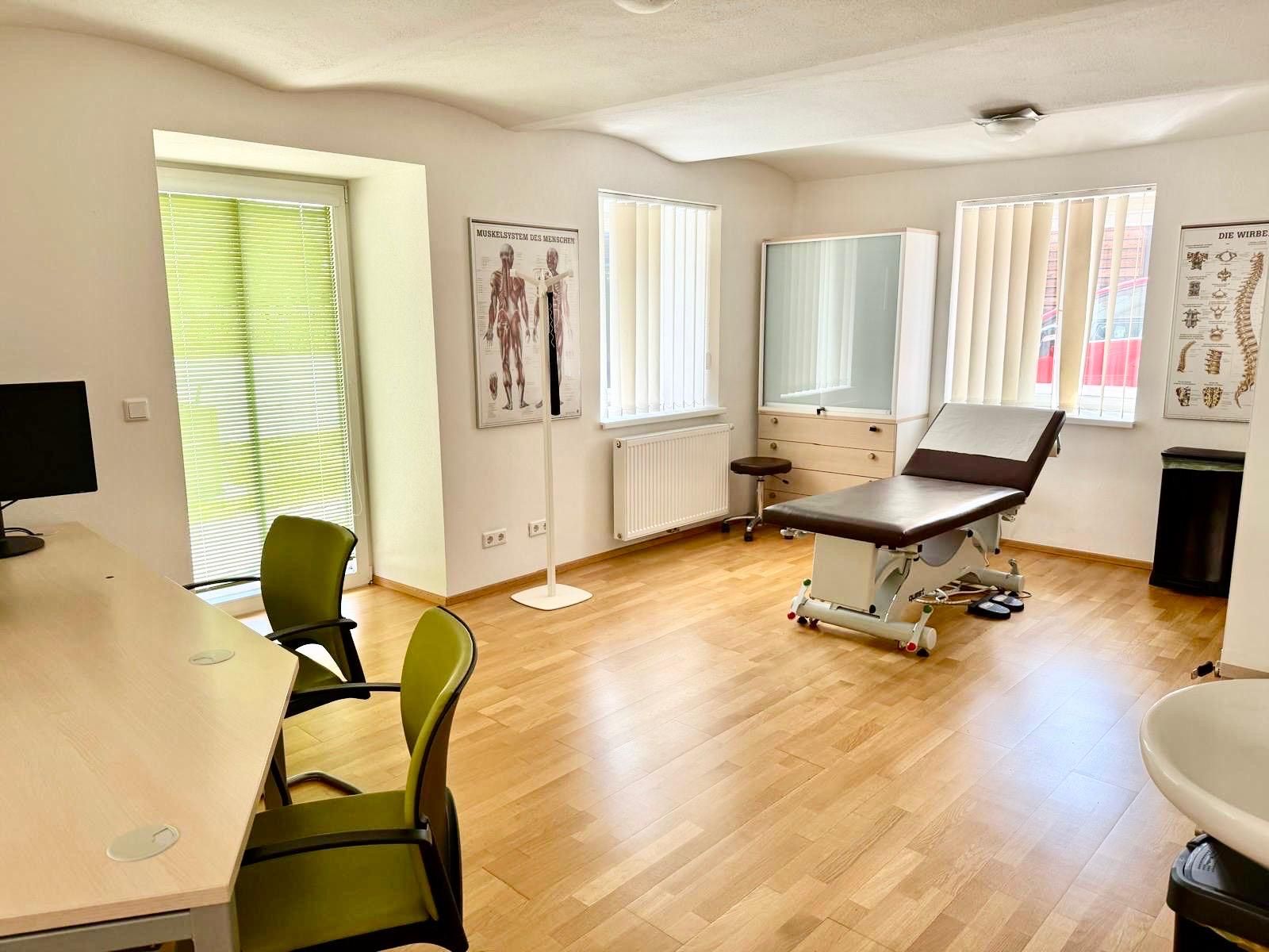 Physiotherapieraum mit Untersuchungstisch, Schreibtisch, anatomischen Diagrammen und zwei grünen Stühlen.