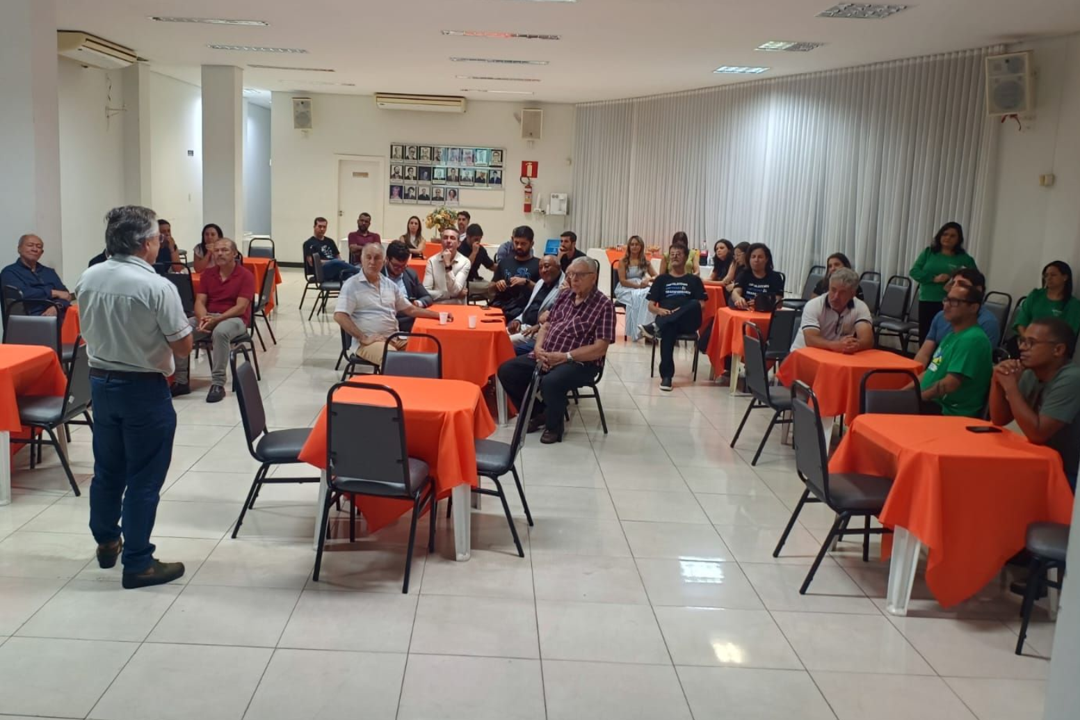 A man speaks to a seated audience in a room with orange tablecloths.