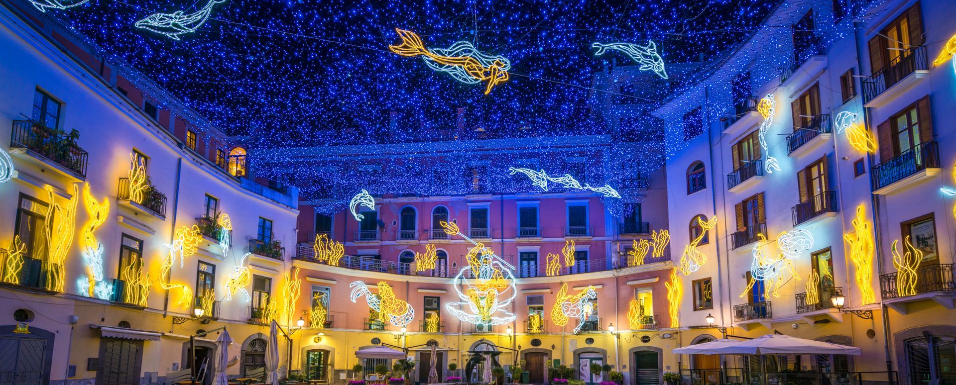 Piazza festosa di notte con luci blu a forma di stelle sopra la testa ed edifici e decorazioni illuminati.
