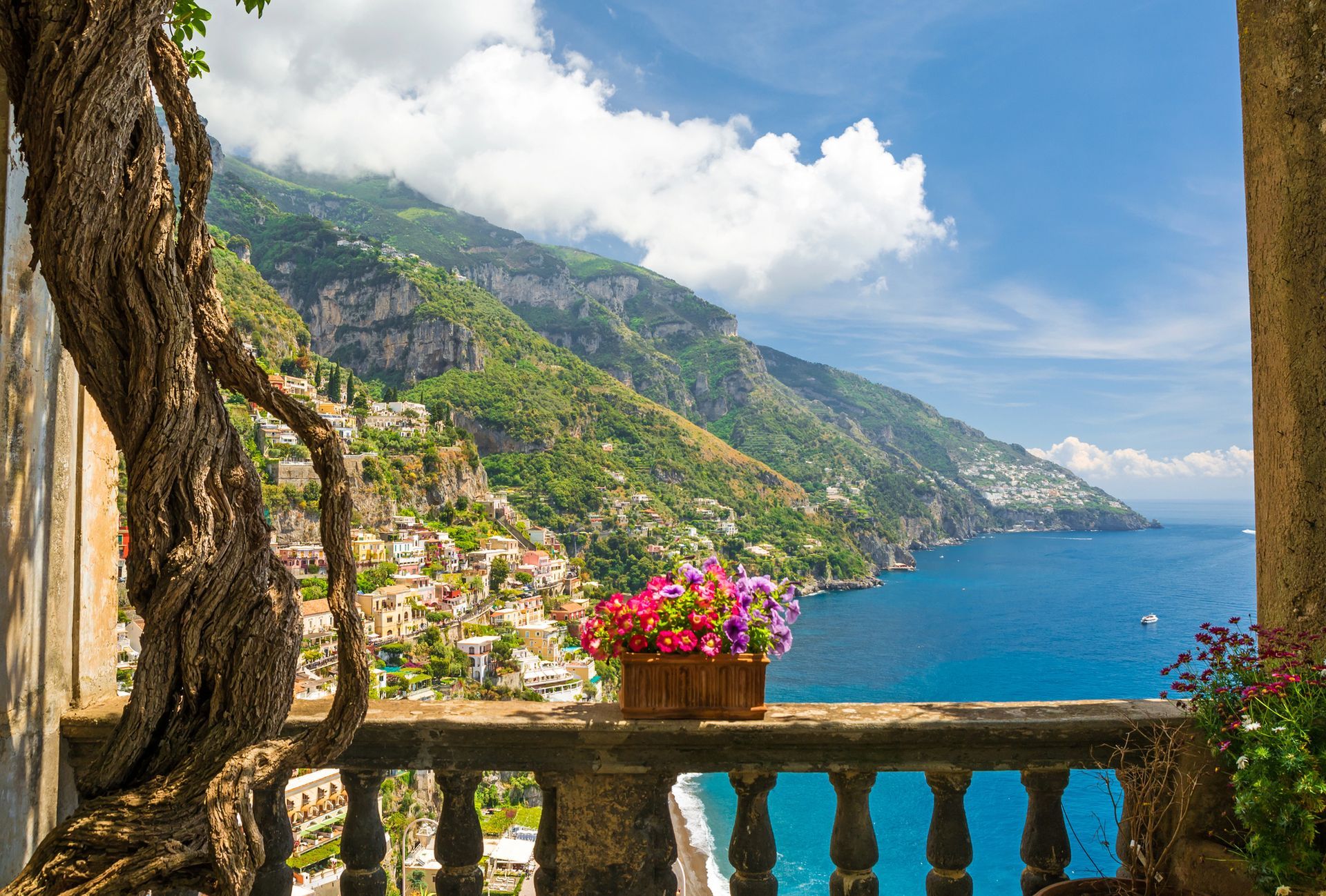 Splendida vista costiera da un balcone fiorito, con vista su una baia azzurra e un villaggio collinare sotto un cielo luminoso.