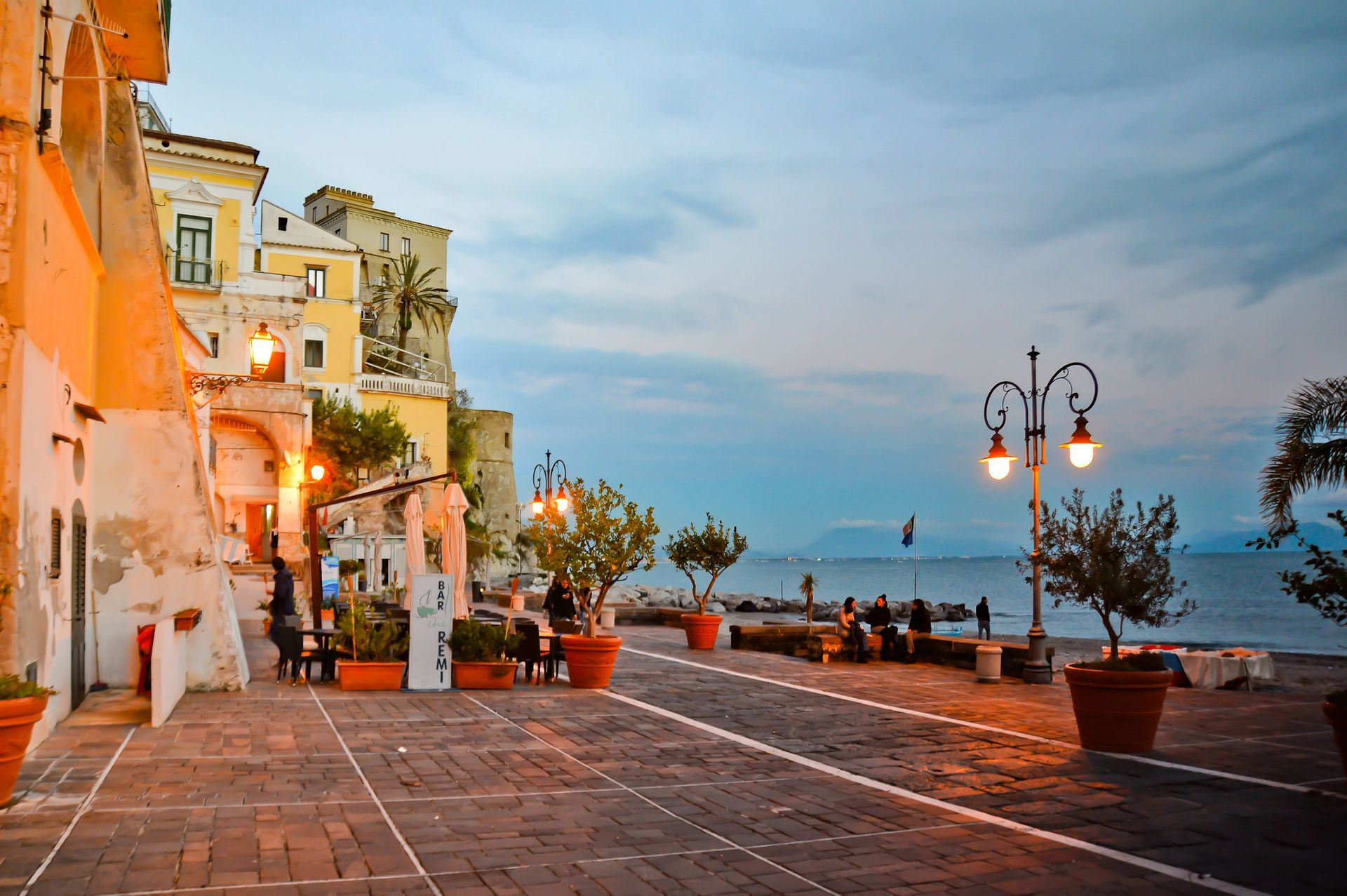 Passeggiata costiera al crepuscolo con edifici colorati, lampioni, piante in vaso e vista sul mare.