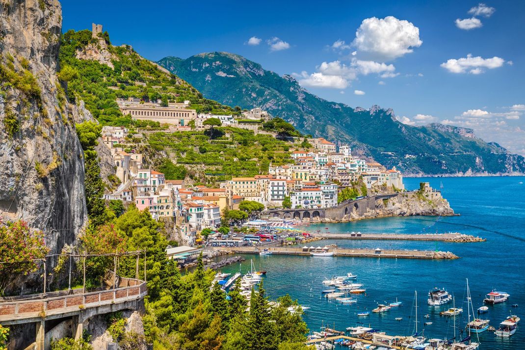 Colorato villaggio costiero arroccato su una scogliera a picco su acque azzurre, con barche e montagne sotto un cielo limpido.