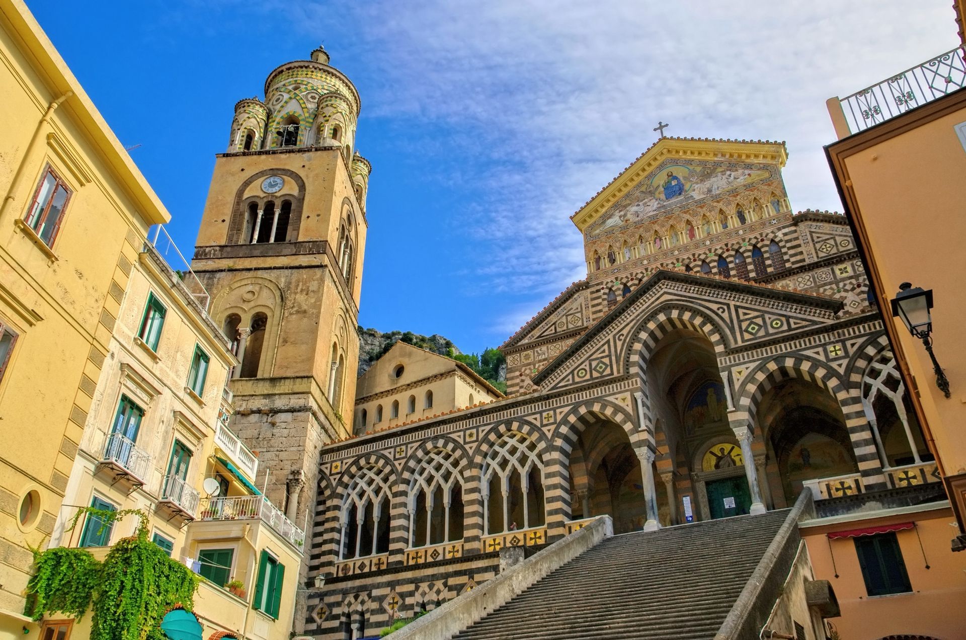 Facciata della cattedrale con archi a strisce e alto campanile su una soleggiata strada in cima a una collina.