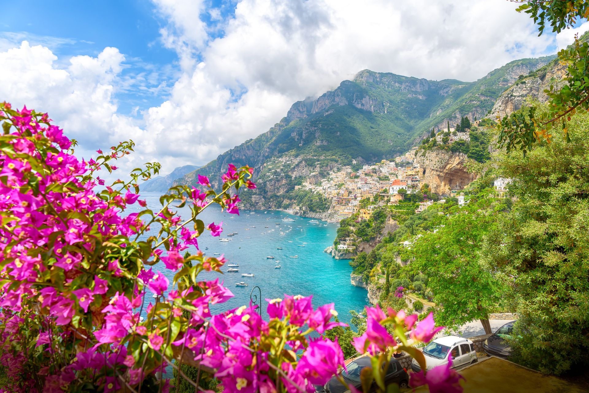 Baia turchese accanto a un colorato villaggio collinare, incorniciata da bouganville rosa acceso e montagne verdi sotto le nuvole.