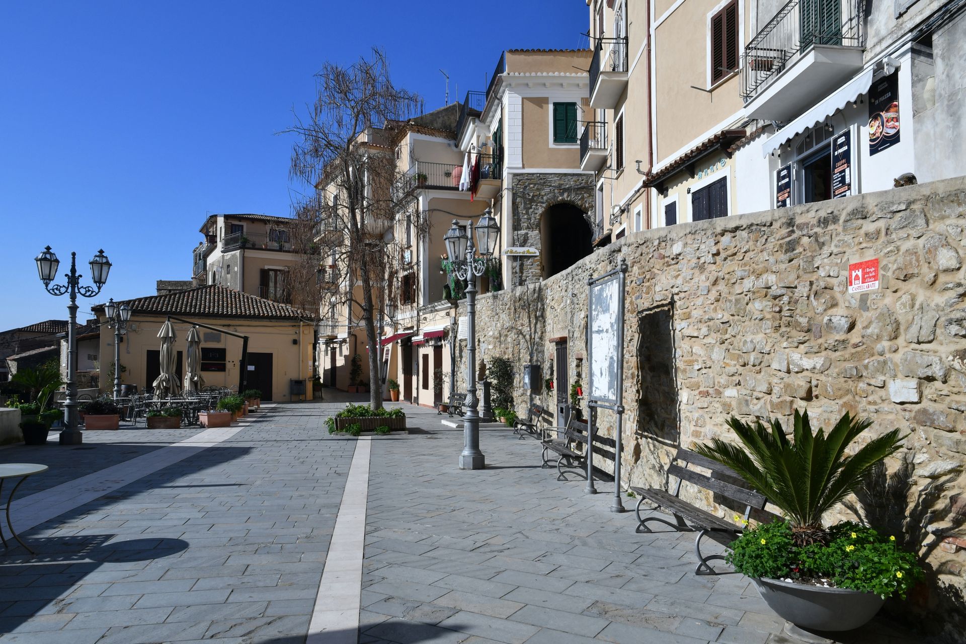 Piazza lastricata in pietra accanto a edifici color sabbia e un muro di pietra sotto un cielo azzurro limpido.