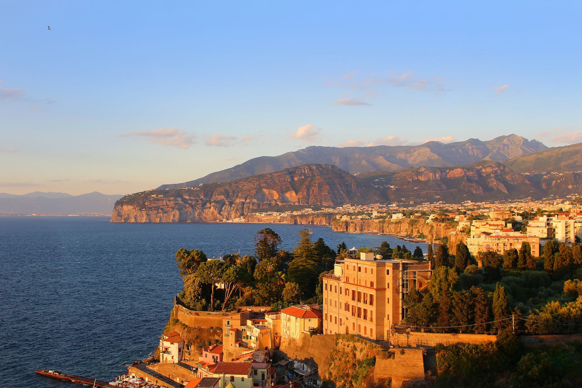 Città costiera al tramonto con litorale roccioso, mare blu e montagne sullo sfondo.