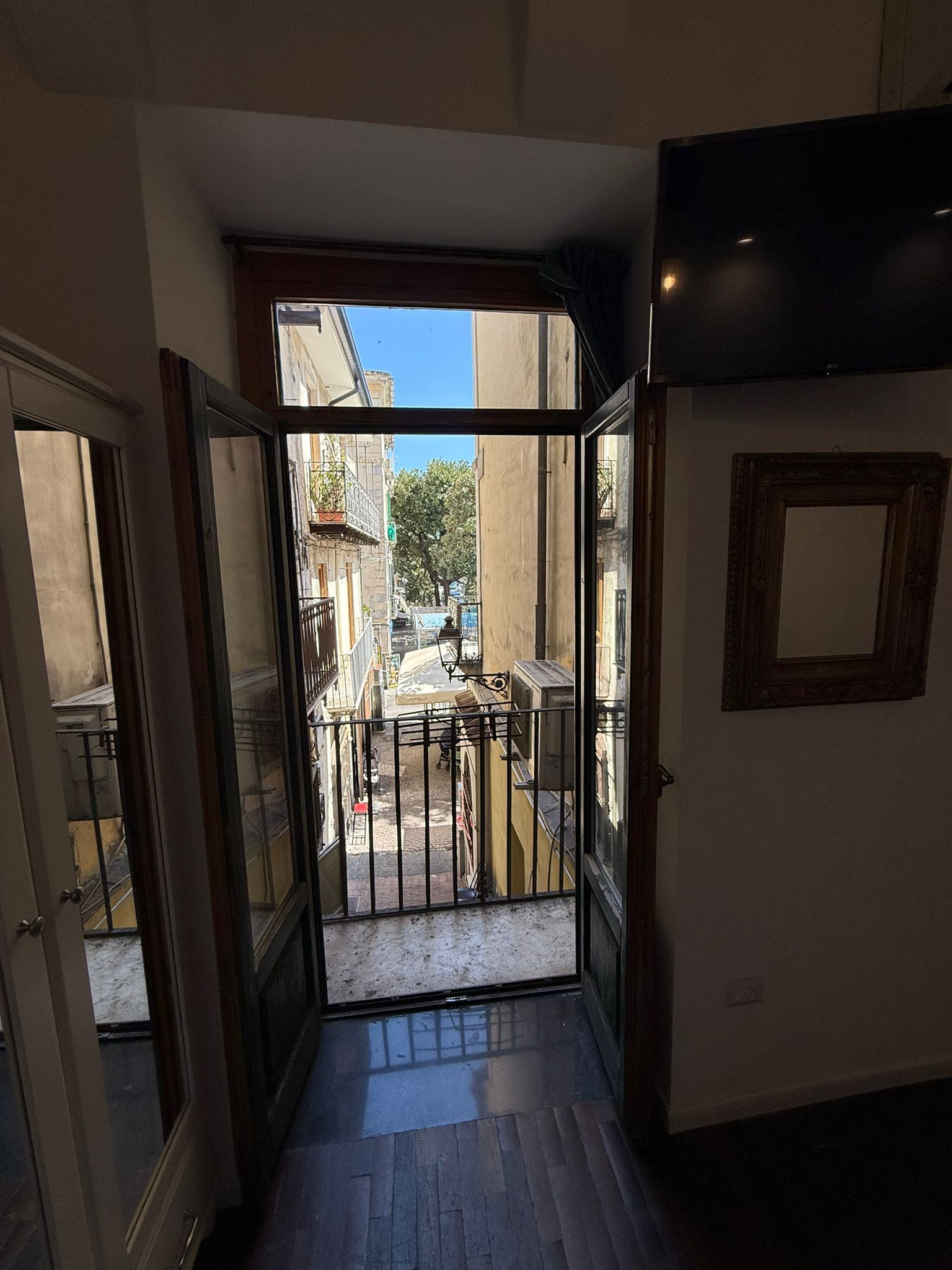 Corridoio che si apre su un balcone soleggiato con ringhiera in ferro e vista su una strada della città.