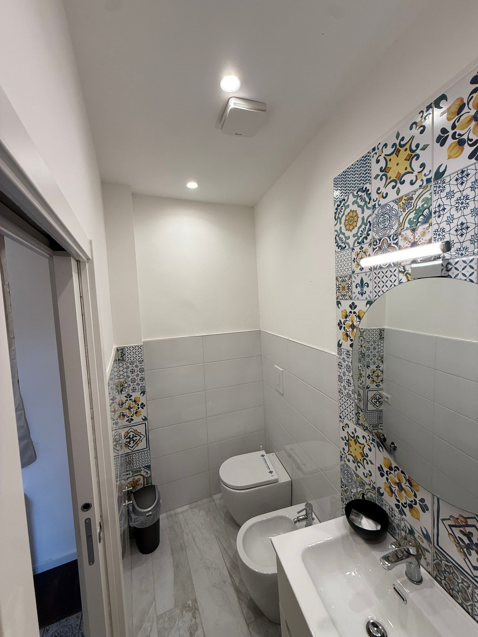 Piccolo bagno piastrellato con WC, lavandino, specchio e una parete decorativa con motivi colorati.