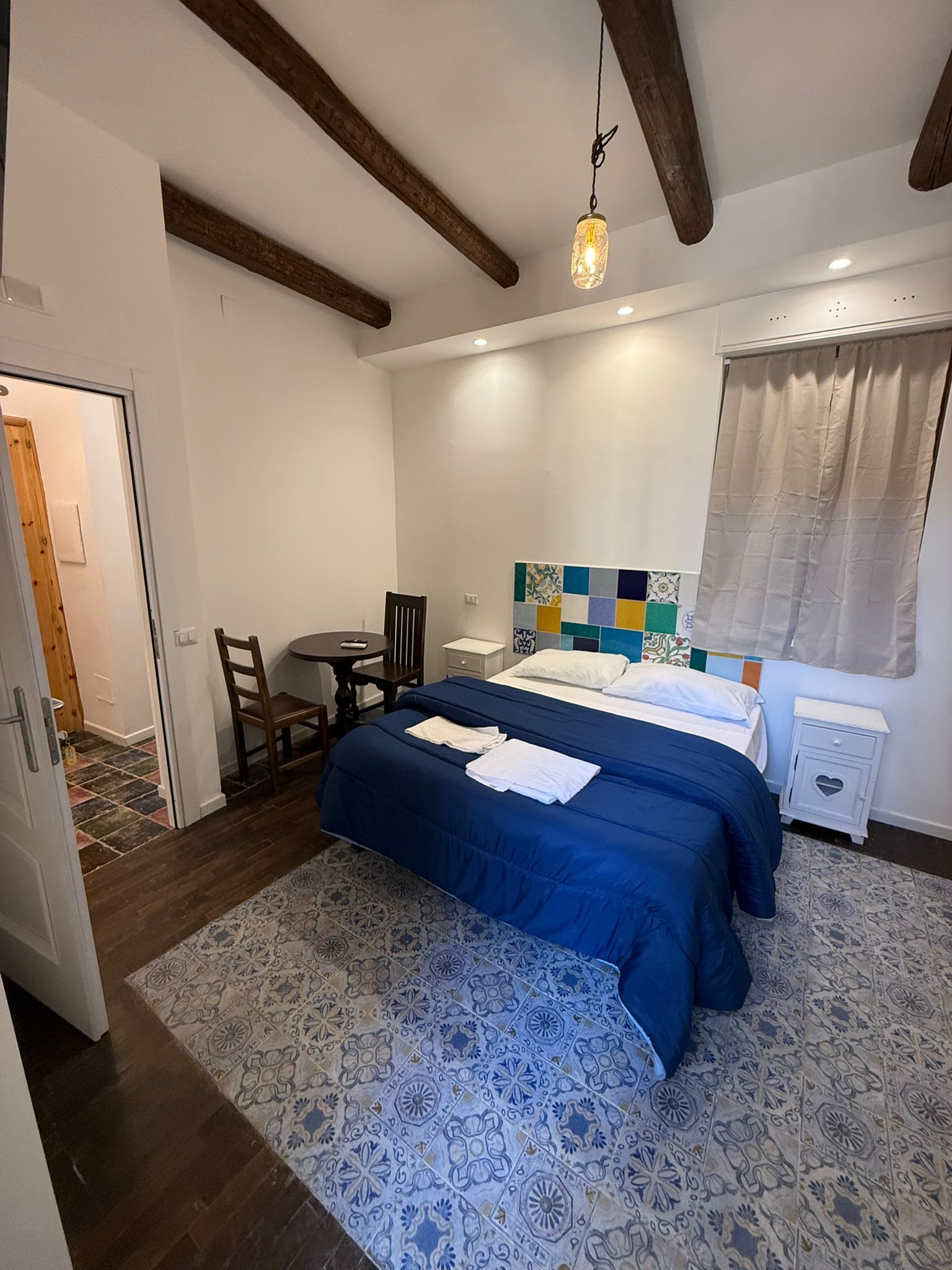 Accogliente camera da letto con copriletto blu, tappeto a motivi geometrici, travi a vista e finestra con tenda.