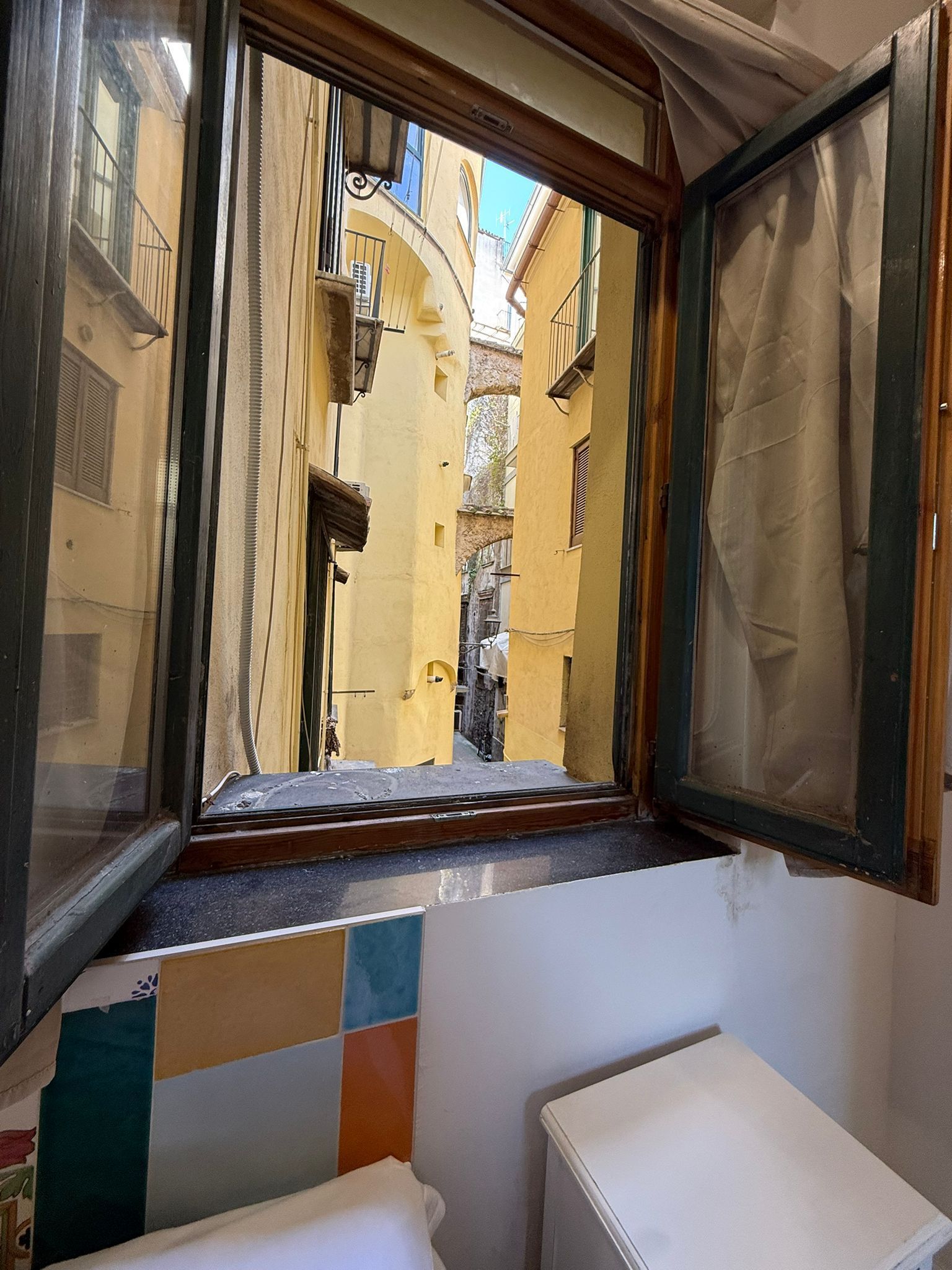 Finestra di legno aperta che si affaccia su stretti edifici e balconi gialli, con piastrelle colorate sottostanti e un WC bianco all'interno.