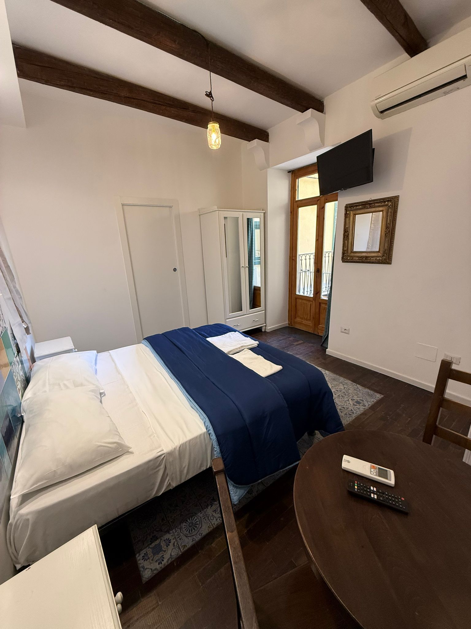Camera da letto luminosa con copriletto blu, armadio bianco, travi in legno e una piccola scrivania vicino alla finestra.