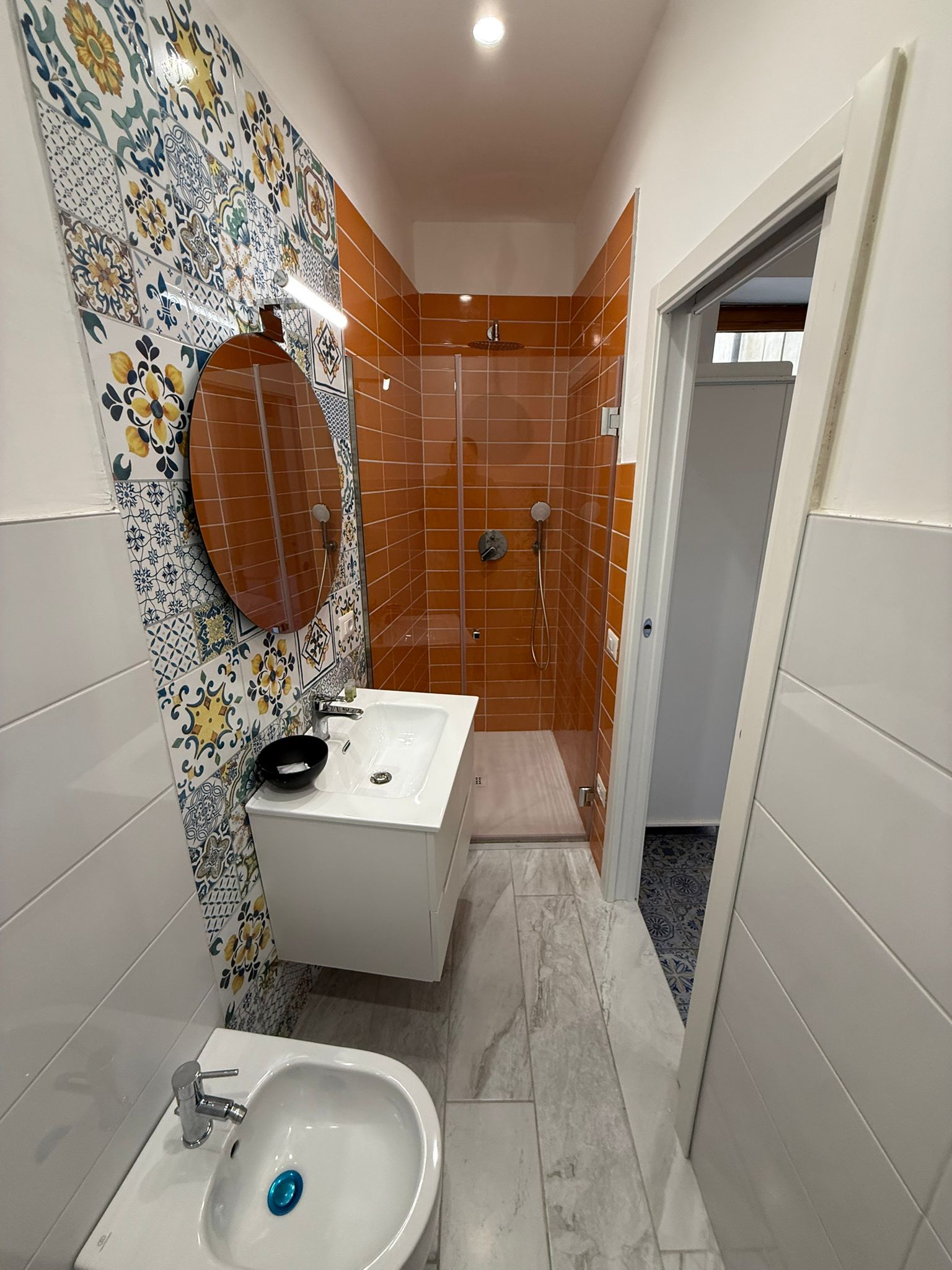 Bagno stretto con parete a mosaico, piastrelle arancioni nella doccia, lavabo, bidet e porta aperta.