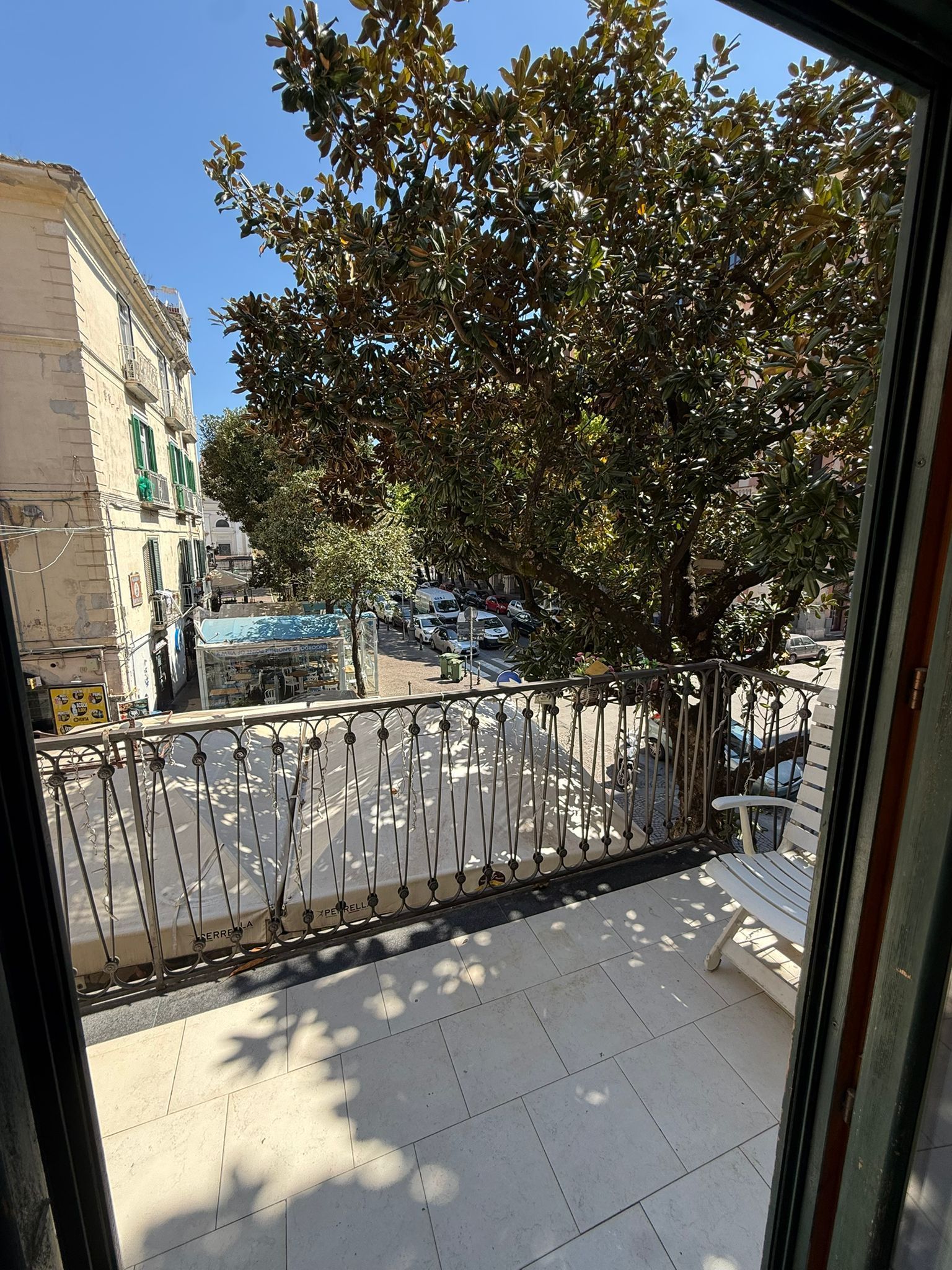 Balcone con pavimento piastrellato, ringhiera in metallo, grande albero e vista sulla strada.