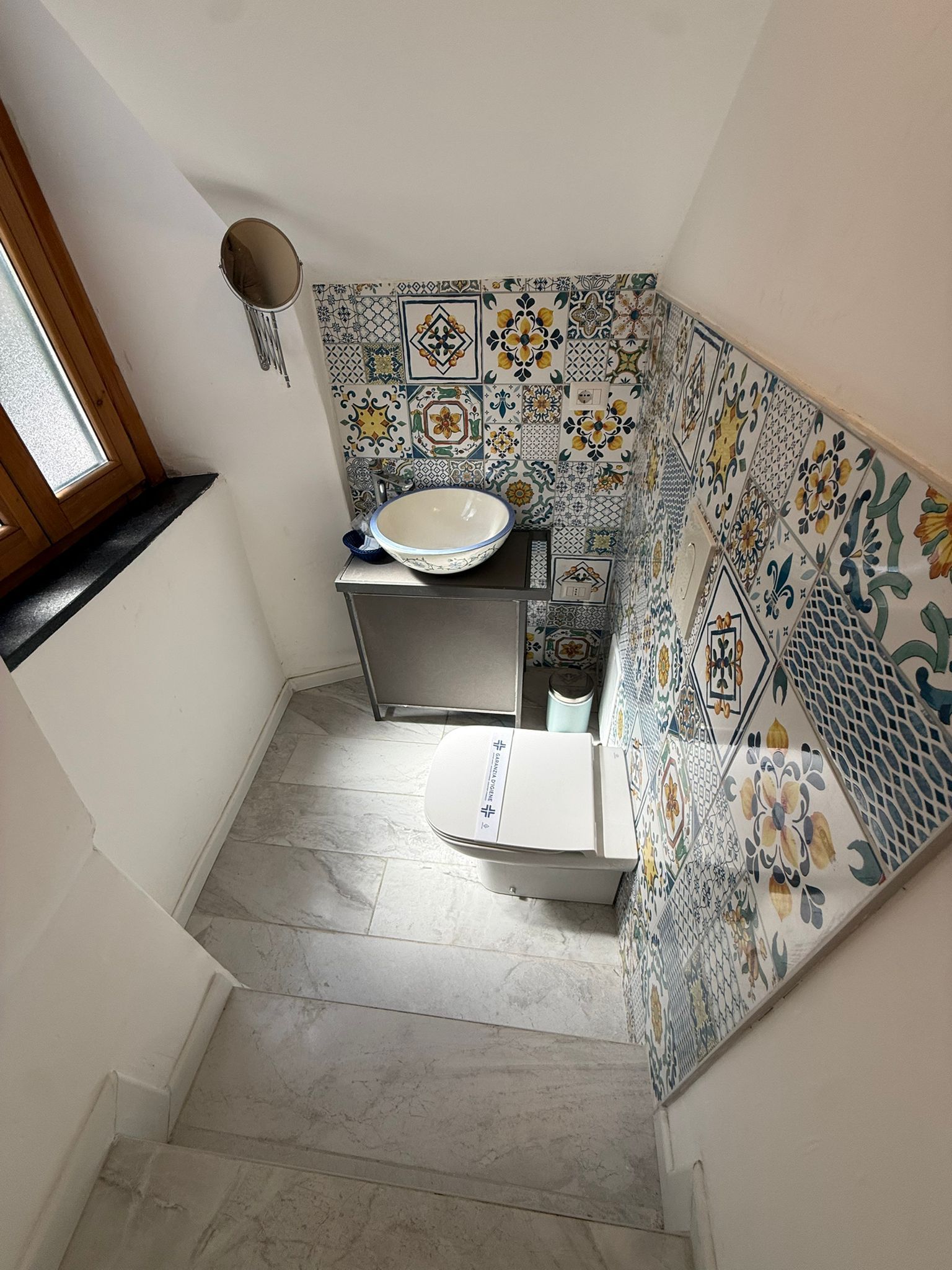 Una stretta scala conduce a un piccolo bagno piastrellato con lavandino, WC e decorazioni a mosaico sulle pareti.