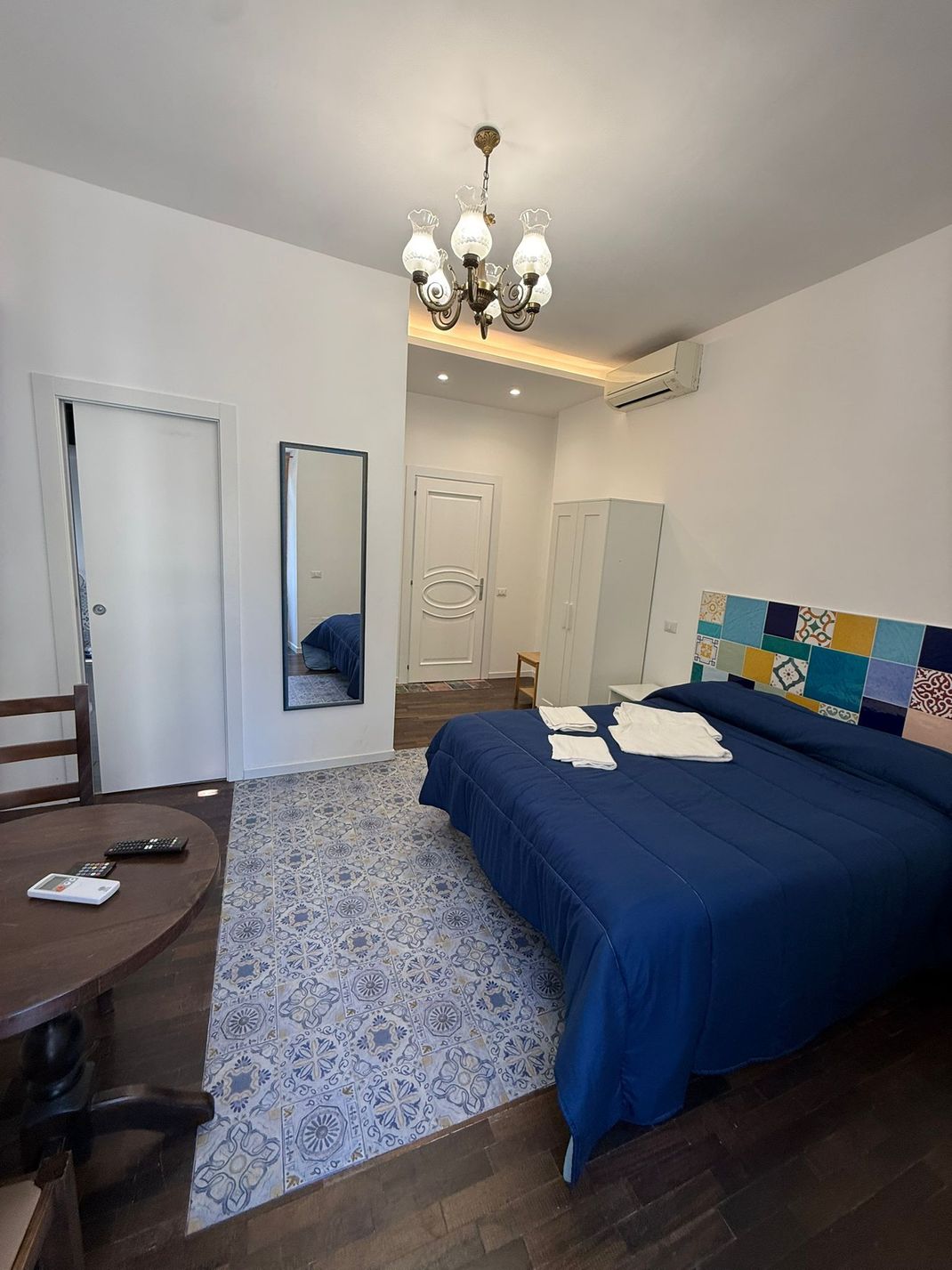 Camera da letto con copriletto blu, tappeto a motivi geometrici, toletta e lampadario in una stanza luminosa e bianca.