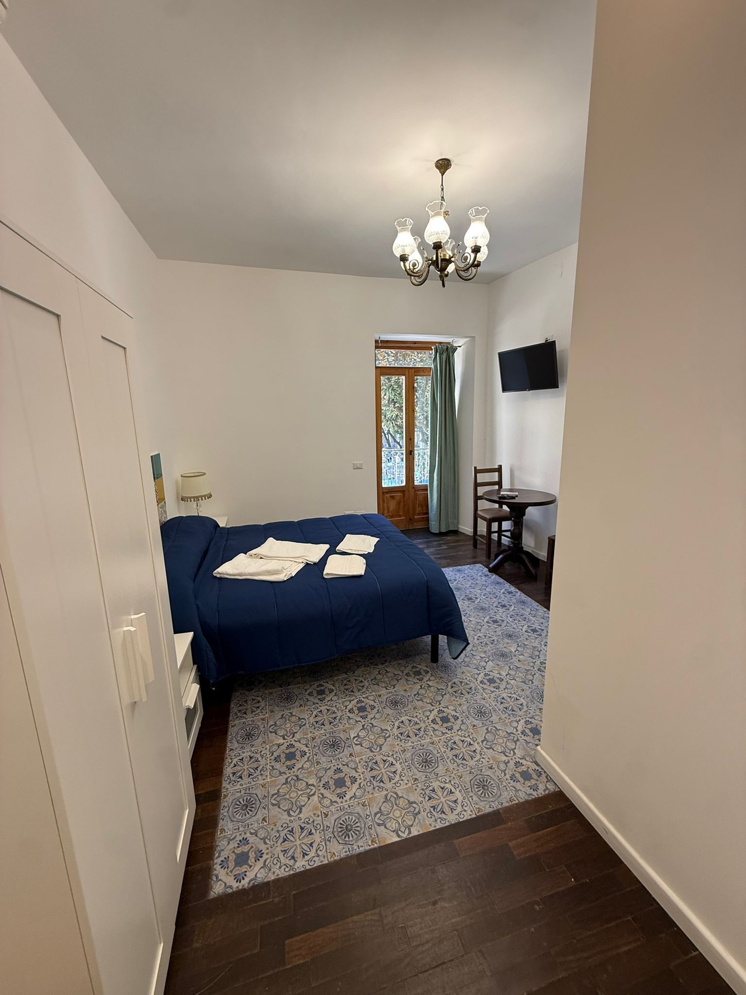 Camera da letto con copriletto blu, tappeto a motivi geometrici, TV a parete e lampadario vicino a una finestra.