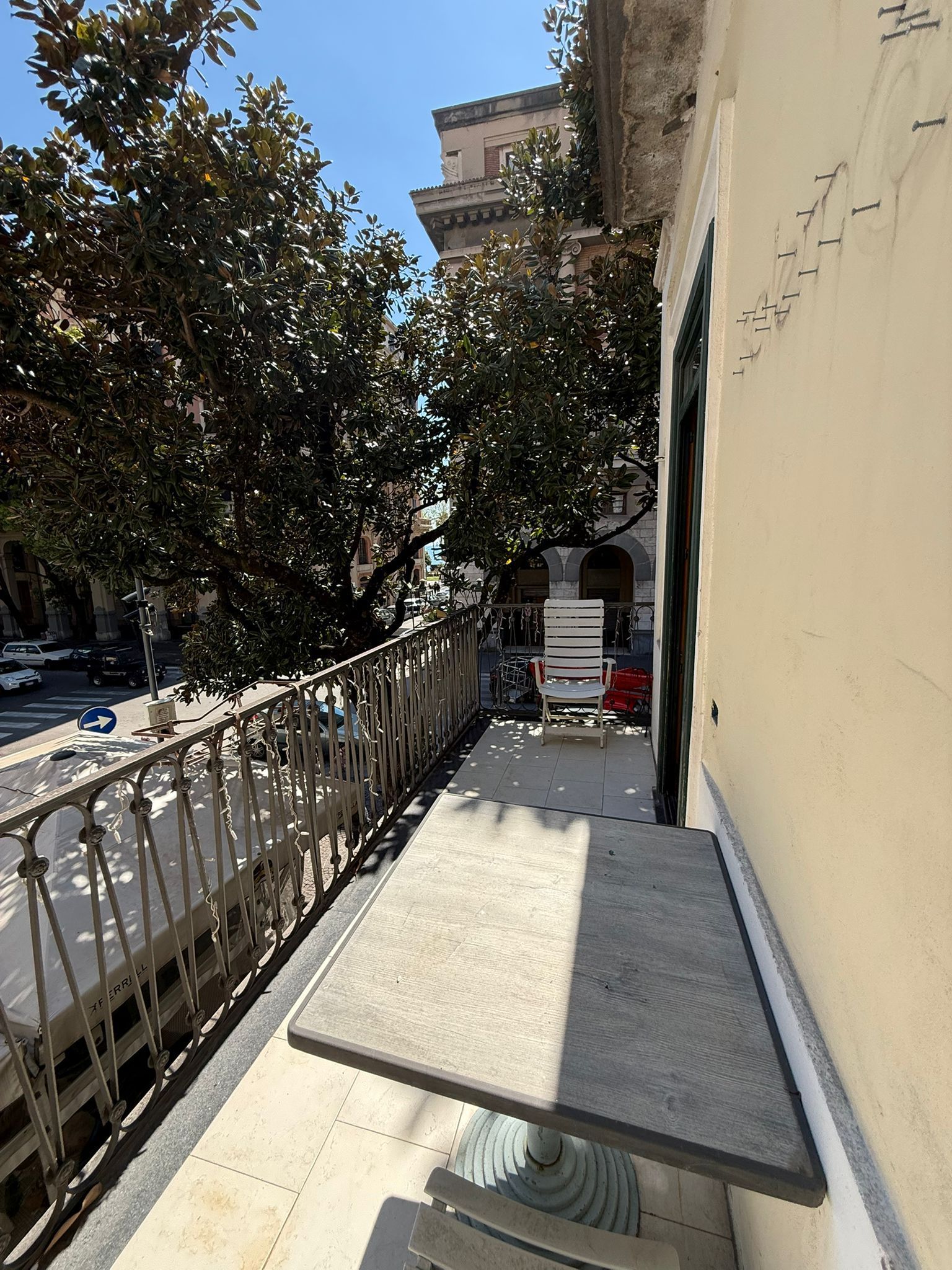 Stretto marciapiede accanto a un muro beige con ringhiera, alberi e una luminosa vista su una strada della città.