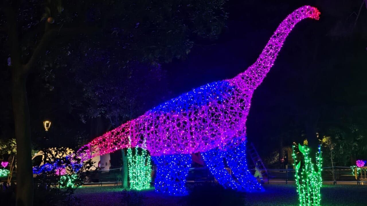 Grande scultura luminosa a forma di dinosauro che brilla di rosa, viola e blu in un'installazione notturna all'aperto.