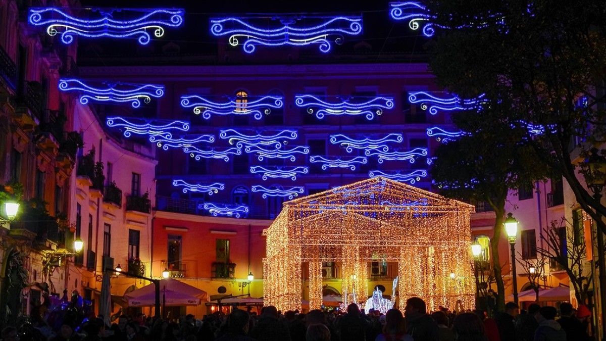 Una strada notturna addobbata con luci natalizie blu e oro, con una tettoia luminosa e una folla sottostante.