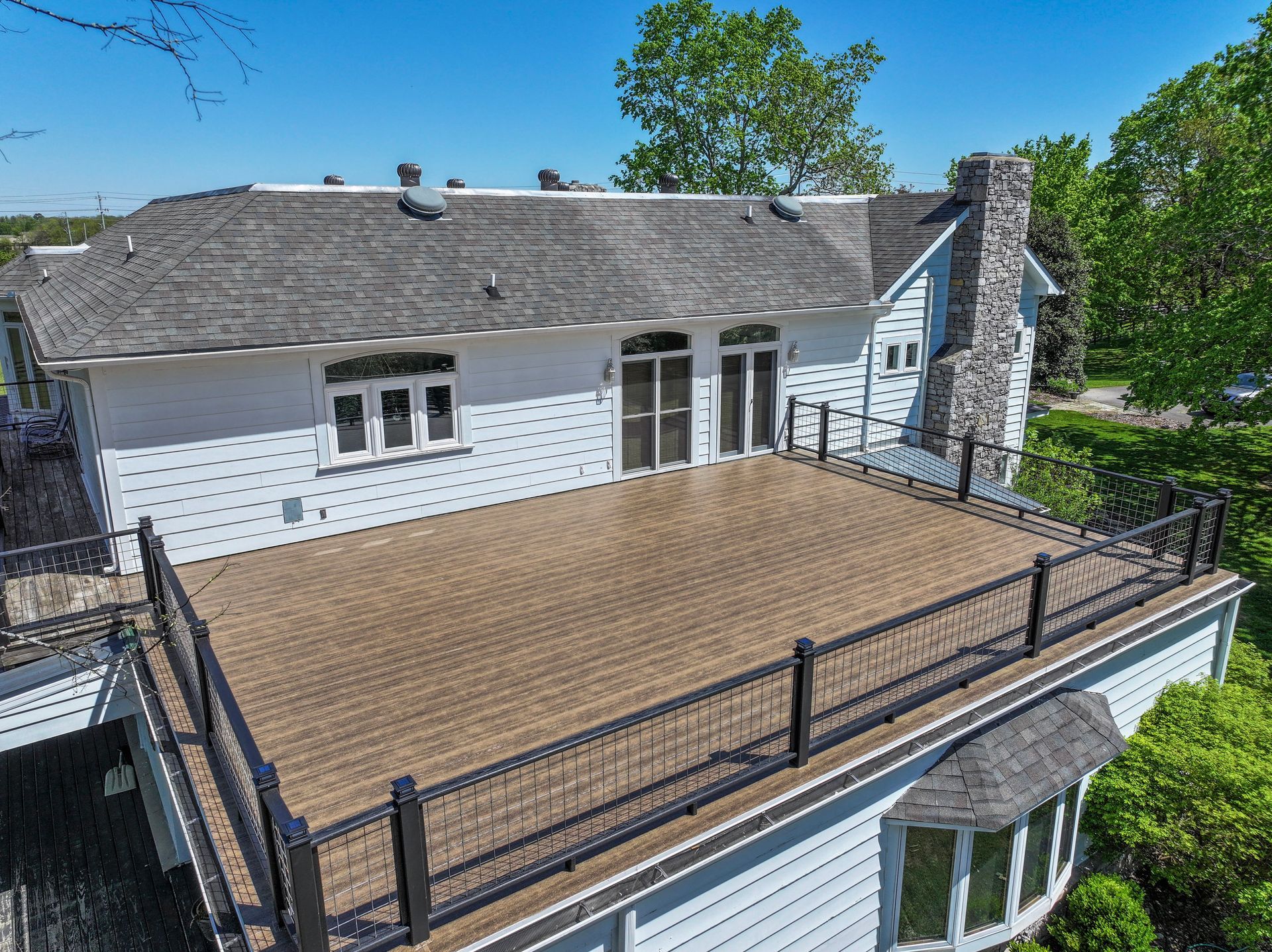 White house with large deck, black railing, and stone chimney. Sunny day.