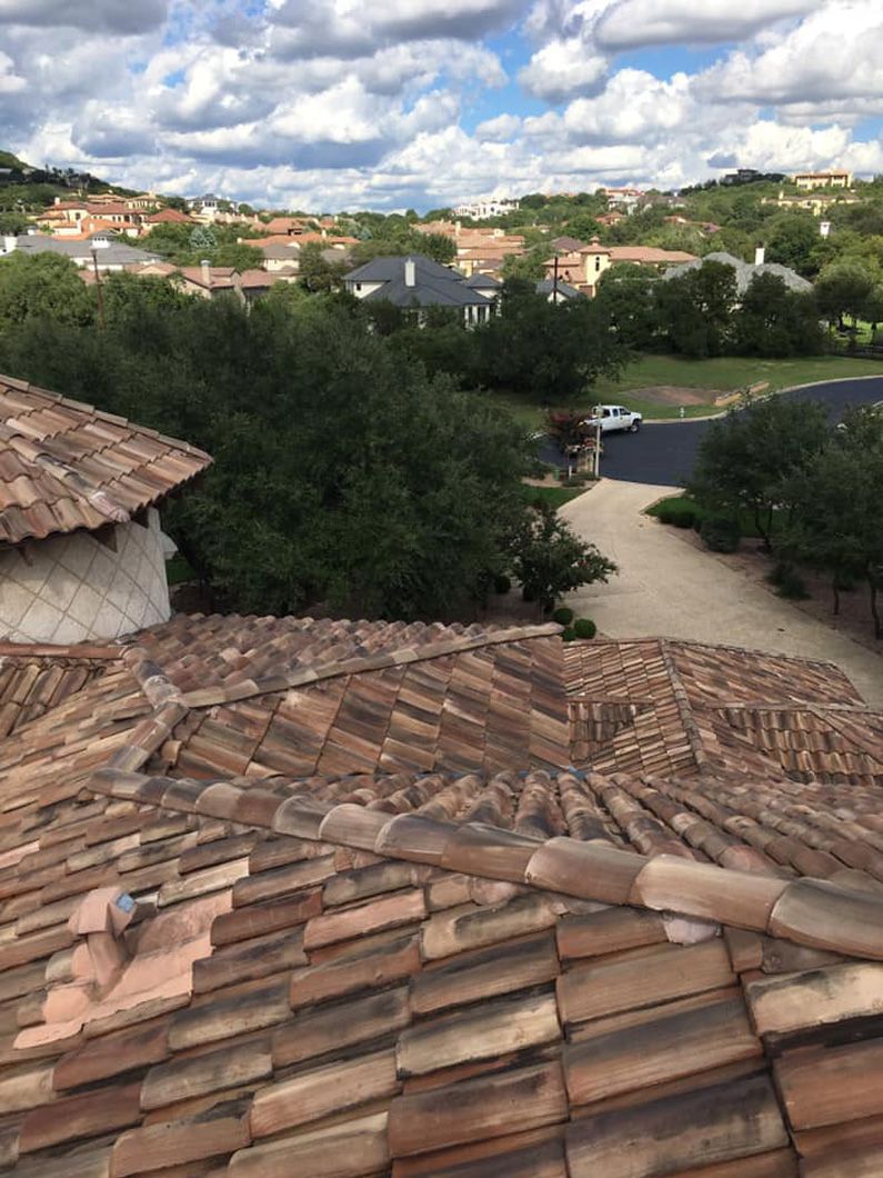Rooftop view of a neighborhood with clay tile roofs, trees, winding road, and partly cloudy sky.