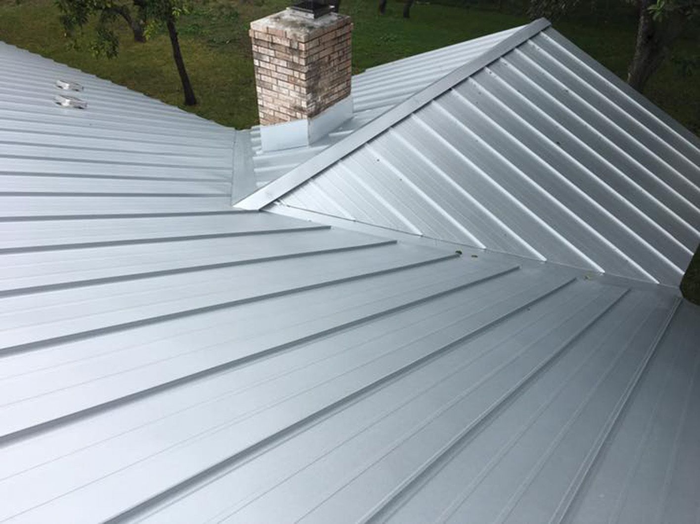 Silver metal roof on a house, with a brick chimney in the center. Green grass in the background.