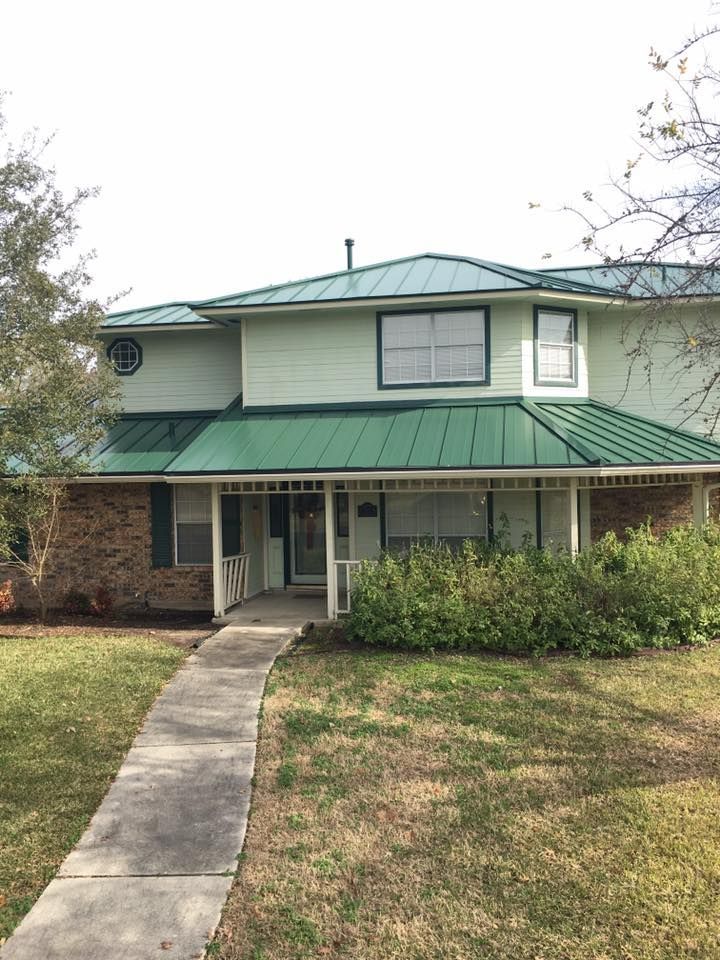 Two-story house with green metal roof, brick base, and concrete walkway leading to the front door.