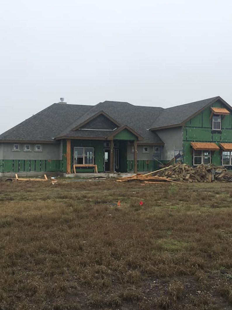 House under construction on a grassy field; gray siding, green sheathing, wood framing visible.