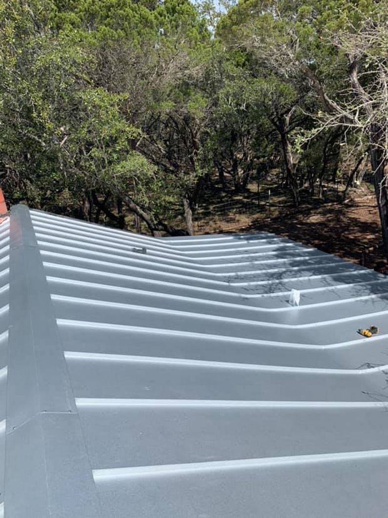 Metal roof with horizontal ridges, painted light gray, set amongst trees.