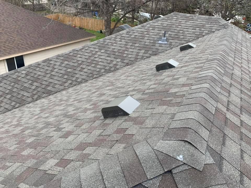 Close-up of a shingled roof with three silver vents. The shingles are various shades of brown and gray.